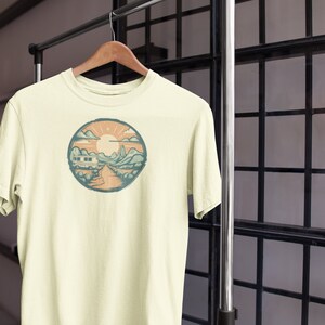 May include: A cream-coloured t-shirt with a circular graphic of a camper van driving through a mountain landscape with a sun and clouds in the sky.