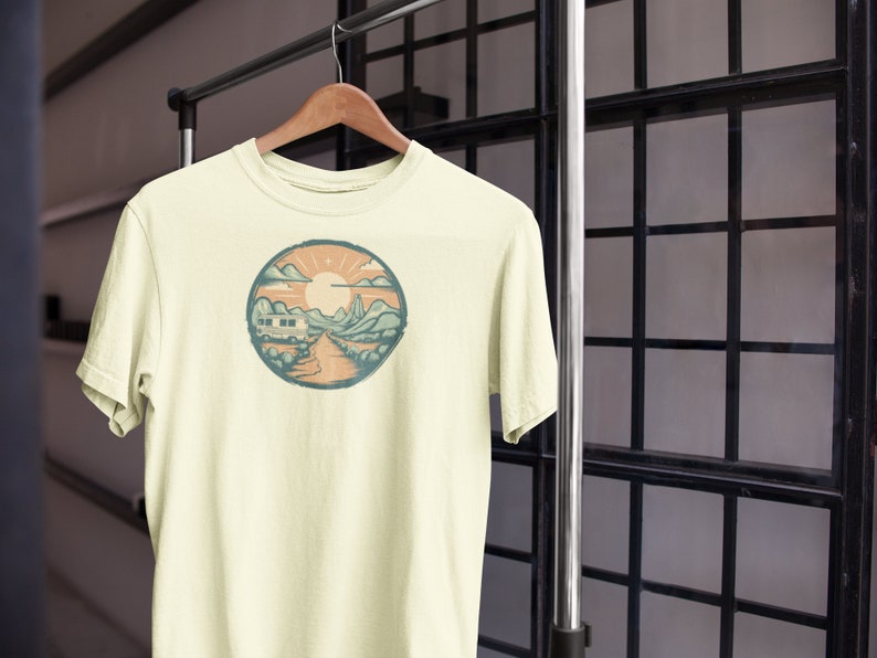 May include: A cream-coloured t-shirt with a circular graphic of a camper van driving through a mountain landscape with a sun and clouds in the sky.