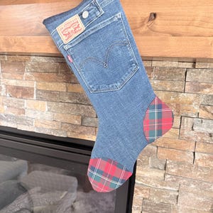 May include: A Christmas stocking made from blue denim, featuring a Levi Strauss & Co. label and a red plaid pattern on the toe and heel. The stocking is hanging from a wooden mantel.