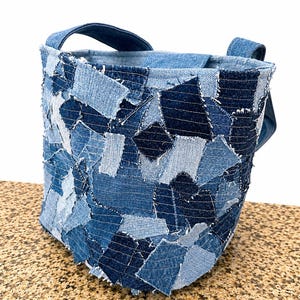May include: A bucket-style bag constructed from a patchwork of various denim fabric scraps in multiple shades of blue. The exterior features overlapping rectangular and irregular denim pieces with visible stitching, creating a textured surface with two attached handles.