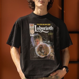 Pode incluir: Uma pessoa vestindo uma camiseta preta com um gráfico de uma capa de quadrinhos com o texto "Labyrinth" e um personagem do filme. A pessoa está vestindo calças camufladas e óculos de sol.
