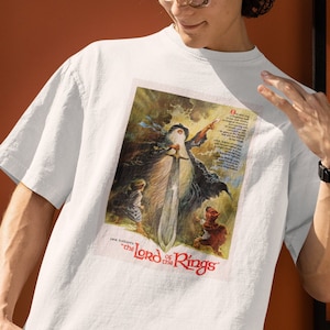 Op de afbeelding: Wit T-shirt met een vintage illustratie van Gandalf uit "The Lord of the Rings" die een zwaard vasthoudt. De illustratie is in een kleurrijke stijl met een donkerblauwe achtergrond.