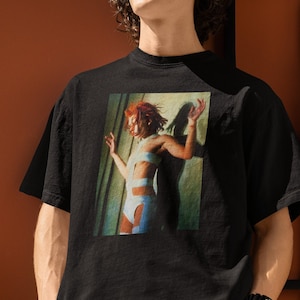 May include: A black t-shirt with a graphic print of a person in a blue and white outfit. The person is standing in front of a window with green curtains.