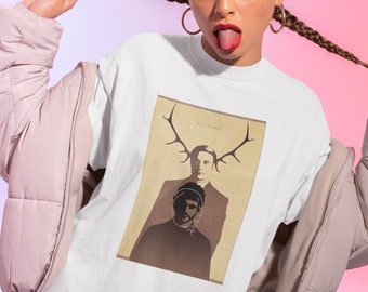 May include: White t-shirt featuring a graphic print of a man with antlers. The graphic is in brown and beige tones. The text "Hannibal" is printed above the graphic.