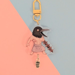 May include: A clear acrylic keyring featuring a cartoon warrior with a bird-like head, holding a spiked club. The keyring has a gold-coloured clasp and a small pinecone charm. The background is split diagonally, with pink and blue sections.