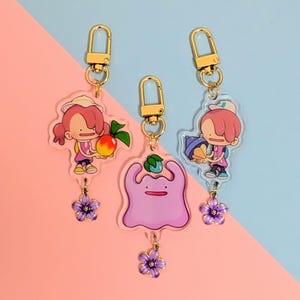 May include: Three acrylic keychains with gold-colored hardware. The keychains feature cartoon characters and a purple Ditto. Each keychain has a dangling purple flower charm. The background is split between pink and blue.