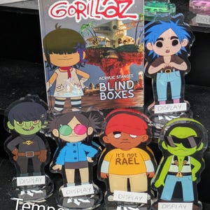May include: A Gorillaz acrylic standee blind box packaging featuring a cartoon character illustration, alongside five distinct character standees on clear circular bases. Each standee is labeled with a white display tag at the base.