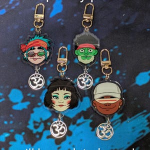 May include: Four acrylic keychains featuring cartoon character heads with gold-tone lobster clasps and small silver-tone Om symbol charms. The characters include a green-haired figure, a green-skinned figure, a dark-haired figure, and a tan-skinned figure.