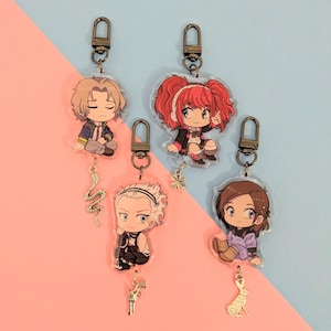 Puede incluir: Cinco llaveros de personajes de estilo anime con herrajes dorados. Cada llavero presenta un diseño de personaje diferente, con dijes colgantes que incluyen una serpiente, un trébol, una pistola y un conejo. El fondo está dividido entre rosa y azul.