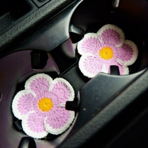 May include: Two crocheted pink flower-shaped cup coasters with white edging and yellow centers are placed in the cup holders of a car.