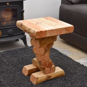 May include: A rustic wooden side table. The table is constructed from stacked, natural-coloured wood logs. The top is a square of wood, and the base is made of two parallel logs. The table is approximately 61 cm tall.