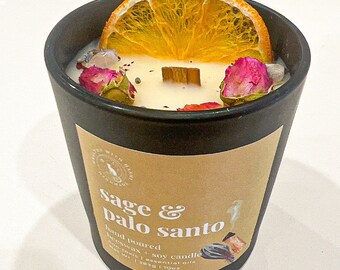 Puede incluir: Un tarro de vela negro con aroma a salvia y palo santo. La vela está cubierta con una rodaja de naranja, capullos de rosa secos y un palo de madera. La etiqueta dice "sage & palo santo hand poured beeswax + soy candle non-toxic + essential oils net wt. 12oz | 340g"