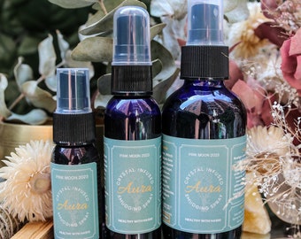 May include: Three glass bottles with blue liquid and spray tops. The bottles are labeled "Crystal Infused Aura Smudging Spray" with the text "Pink Moon 2023" and "Healthy with Hashi" on the labels.