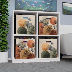 Könnte beinhalten: Ein weißes Würfel-Aufbewahrungsregal mit vier Fächern, jedes mit einer Stoff-Aufbewahrungsbox. Die Boxen haben einen beigefarbenen Hintergrund mit abstrakten floralen Designs in Orange und Grün. Das Regal steht auf einem grauen Teppich.