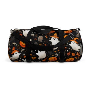 May include: Black duffel bag with a Halloween-themed pattern. The design features white ghosts wearing orange witch hats, pumpkins, and autumn leaves on a black background. The bag has black handles and a shoulder strap.