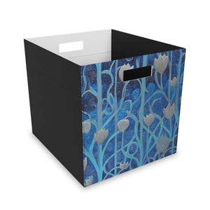 May include: A square storage bin with a black exterior and white interior. The front and sides feature a blue floral design with silver-colored flowers and stems. The bin has two rectangular cut-out handles.