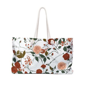 May include: White tote bag with a floral pattern featuring red and peach roses, green leaves, and red berries. The bag has white rope handles.