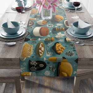 Op de afbeelding: Een turquoise tafelloper met een herhalend patroon van illustraties van eten en drinken. Het ontwerp bevat afbeeldingen van champagneflessen, glazen en diverse etenswaren. De tafel is gedekt met borden, kommen, wijnglazen en een vaas met roze bloemen.