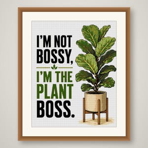May include: A framed wall art print featuring a fiddle leaf fig plant in a light-colored pot on a wooden stand. The text reads: I'M NOT BOSSY, I'M THE PLANT BOSS. The design uses black and green lettering with a small green leaf icon between lines.
