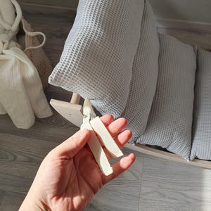 May include: A close-up of a hand holding two white wooden pieces. The pieces are shaped like a long, thin rectangle and are slightly curved. The hand is holding the pieces in front of a light gray and white waffle-textured cushion.