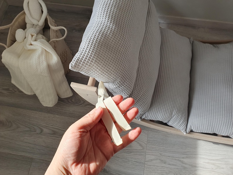 May include: A close-up of a hand holding two white wooden pieces. The pieces are shaped like a long, thin rectangle and are slightly curved. The hand is holding the pieces in front of a light gray and white waffle-textured cushion.