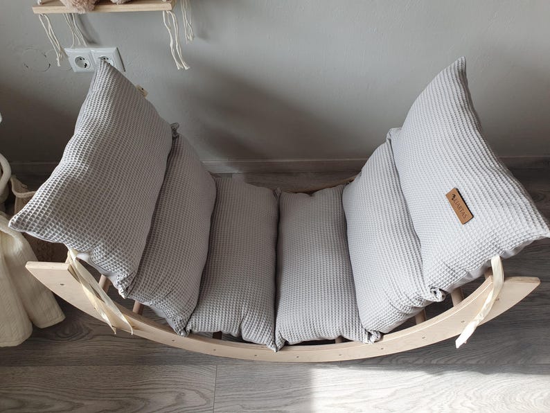 May include: A wooden rocking chair with a gray waffle-textured cushion. The cushion is made up of four separate pieces, each with a different shape. The chair is made of natural wood and has a simple design.