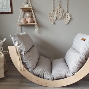 May include: A wooden rocking chair with a gray and white padded cushion. The chair is made of natural wood and has a curved shape. The cushion is made of soft fabric and has a quilted design.