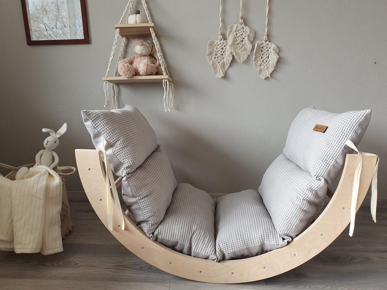 May include: A wooden rocking chair with a gray and white padded cushion. The chair is made of natural wood and has a curved shape. The cushion is made of soft fabric and has a quilted design.