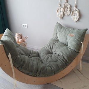 Könnte beinhalten: Ein Schaukelstuhl aus Holz mit einem kissen aus salbeigrünem Cord. Der Stuhl hat eine gebogene Basis und eine gepolsterte Rückenlehne. Der Stuhl ist für Kinder konzipiert und steht auf einem weißen Teppich. Ein Teddybär befindet sich in einem Korb in der Nähe.