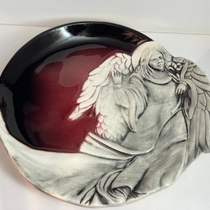 May include: A ceramic dish with a black rim and a burgundy center. The dish features a sculpted angel with wings and a flowing robe.