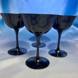 May include: Four black coupe glasses with a rounded bowl and a long stem. The glasses are made of a shiny, dark material and are arranged in a group. The glasses are suitable for serving cocktails or champagne.