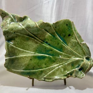 May include: A handcrafted ceramic leaf-shaped dish with a glossy green glaze. The dish features detailed veining and a textured surface, resting on a small metal stand. The color palette includes shades of green and dark accents.