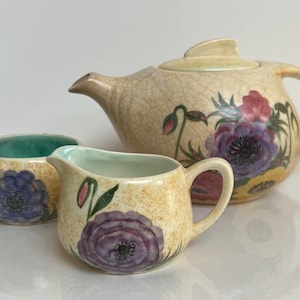 May include: A vintage tea set with a teapot, creamer, and sugar bowl. The set is decorated with a floral pattern in shades of pink, purple, and green on a cream background.