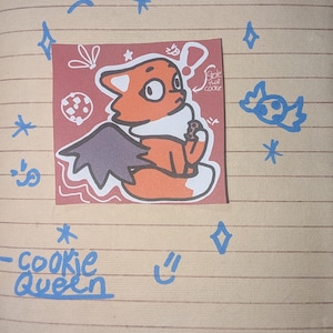 Könnte beinhalten: Ein quadratischer Aufkleber mit einem orangefarbenen fuchsartigen Wesen mit dunklen Flügeln, das einen Keks isst, mit dem Text Stole that cookie und Cookie Queen. Der Aufkleber ist von blauen handgezeichneten Kritzeleien von Sternen und einem geflügelten Keks umgeben.