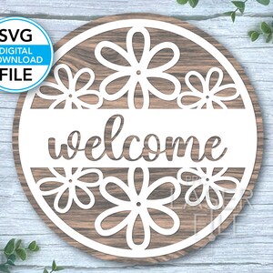 Könnte beinhalten: Ein rundes Holzschild mit einem weißen Holzmaserungsmuster. Das Schild hat einen weißen Ausschnitt des Wortes "welcome" und mehrere stilisierte Blumendesigns.