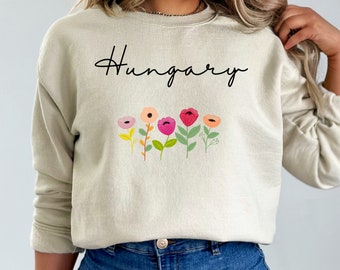 Puede incluir: Una sudadera de color beige claro con la palabra "Hungría" impresa en letra cursiva negra. Debajo del texto se imprime una fila de cinco flores de colores.