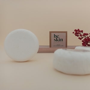 Puede incluir: Una foto de producto con esponjas cosméticas blancas y redondas y una pequeña caja cuadrada con el texto "be skin COSMETICS". Una ramita de bayas rojas añade un toque de color. El fondo es de un tono suave y neutro.