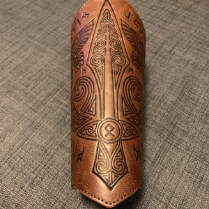 May include: A brown leather arm guard featuring detailed embossed patterns. The design incorporates a central arrow-like element, runes, and swirling motifs. The leather has a textured finish, evoking a medieval or fantasy aesthetic. The arm guard is approximately 20 cm in length.