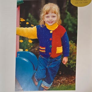 May include: A child wearing a colorful knitted jacket with yellow, blue, red, and white accents. The jacket has a button closure and is made with a textured yarn. The child is standing on a blue playground slide.
