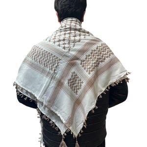 May include: A white and brown checkered keffiyeh scarf with decorative tassels. The scarf is draped over a person's shoulders, showcasing the traditional pattern and fringe detail. The scarf is made of a soft material.