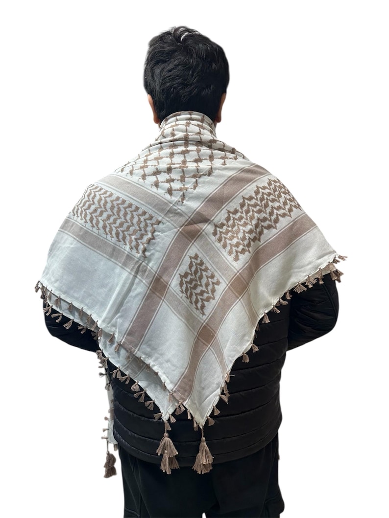 May include: A white and brown checkered keffiyeh scarf with decorative tassels. The scarf is draped over a person's shoulders, showcasing the traditional pattern and fringe detail. The scarf is made of a soft material.