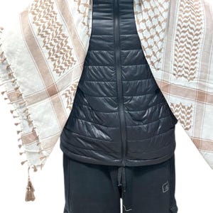 May include: A white and tan keffiyeh scarf with a geometric pattern and tassels. The scarf is draped over a black puffer vest and black pants. The scarf's design features a repeating pattern of squares and lines.