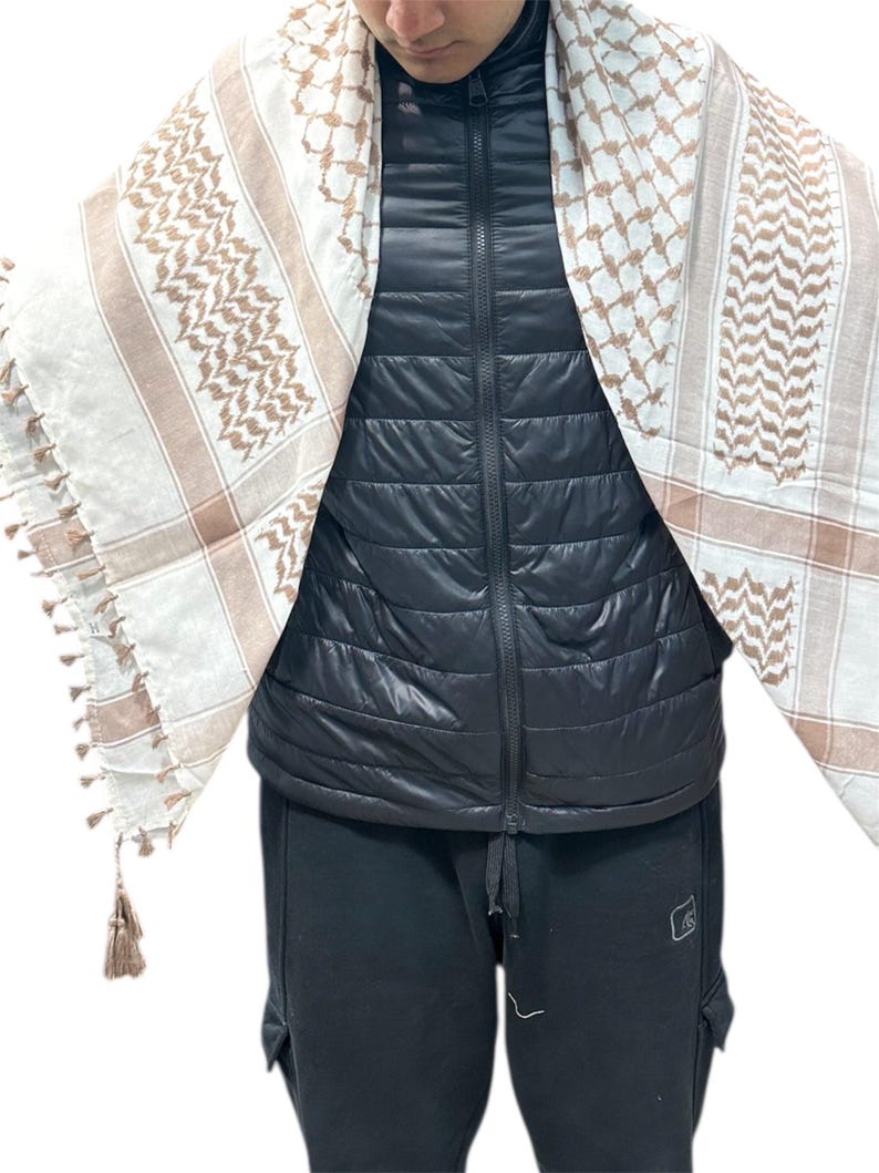 May include: A white and tan keffiyeh scarf with a geometric pattern and tassels. The scarf is draped over a black puffer vest and black pants. The scarf's design features a repeating pattern of squares and lines.