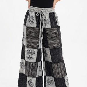 May include: Wide-leg pants featuring a patchwork design in shades of gray and black. The fabric displays alternating squares with printed motifs including suns, moons, trees, and geometric patterns. The pants have an elastic waistband with a drawstring.