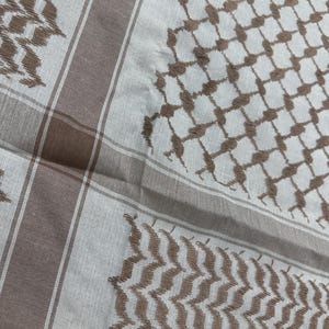 May include: A close-up of a white and brown keffiyeh scarf. The scarf features a repeating geometric pattern in brown, with brown borders and stripes. The fabric is soft and appears to be made of cotton or a similar material.