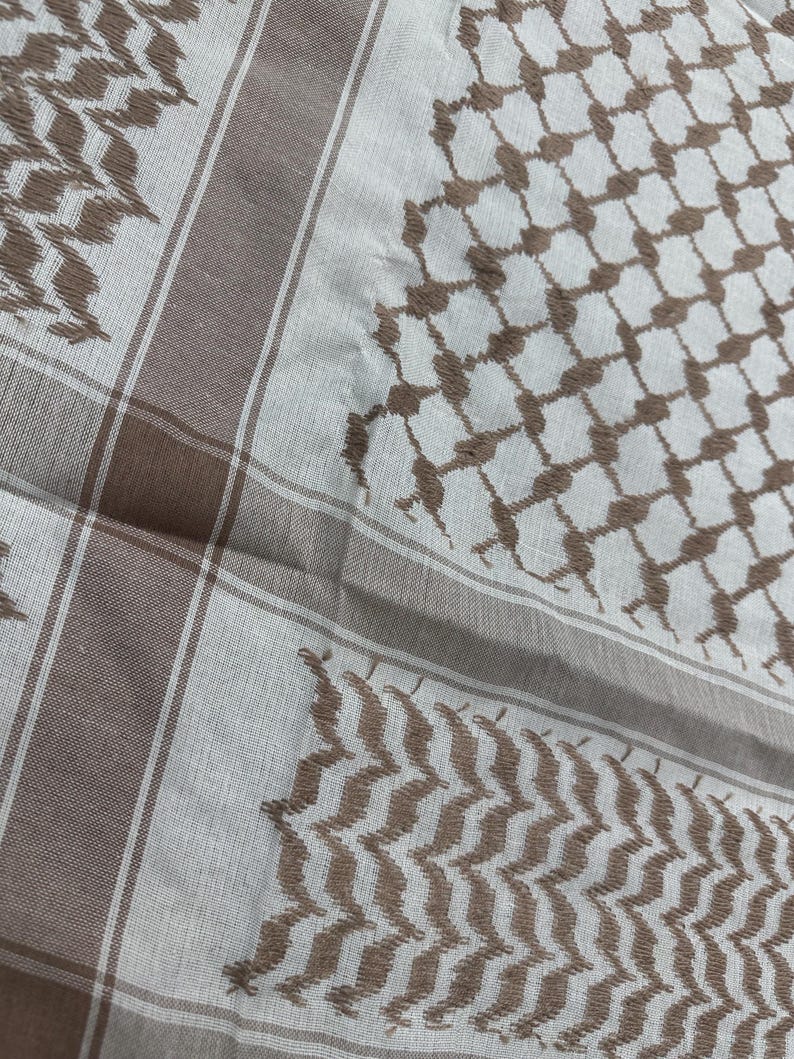 May include: A close-up of a white and brown keffiyeh scarf. The scarf features a repeating geometric pattern in brown, with brown borders and stripes. The fabric is soft and appears to be made of cotton or a similar material.