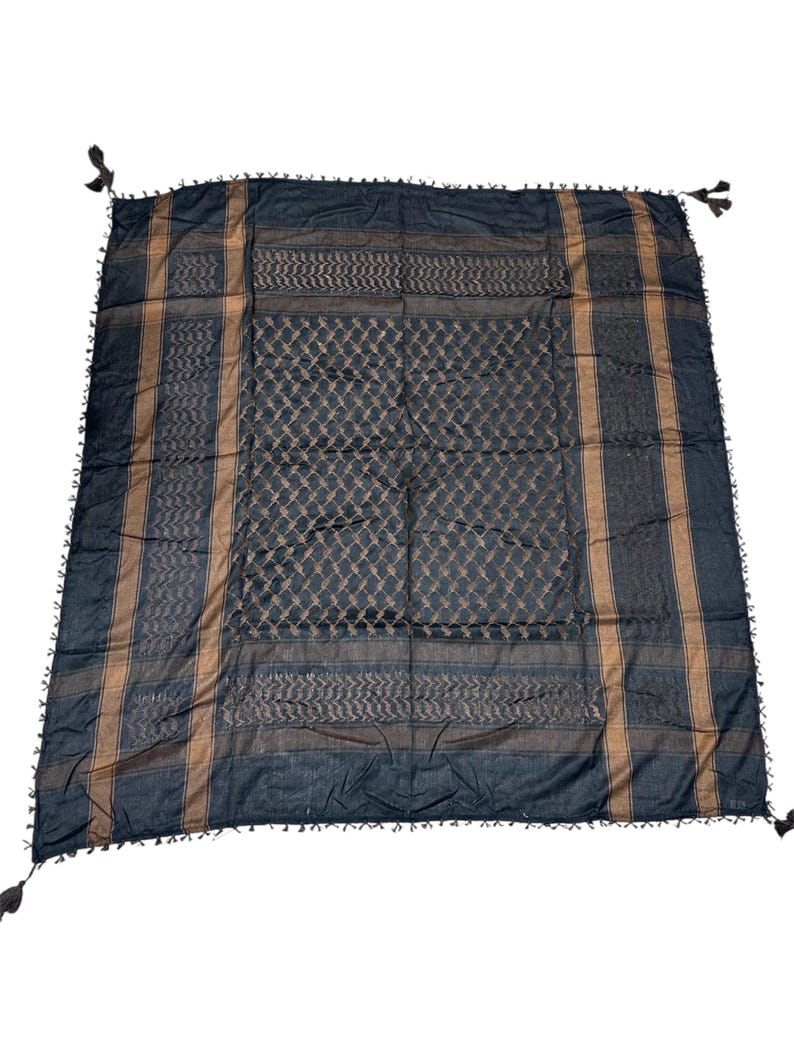 May include: A black and tan keffiyeh scarf with a geometric pattern. The scarf has a diamond and striped design, with tassels on each corner. The scarf is made of a soft material.