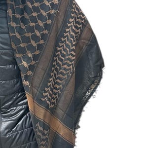 May include: A black and brown keffiyeh scarf with geometric and chevron patterns. The scarf has a fringed edge and is draped over a black jacket. The scarf's design features a combination of dark and light brown hues.