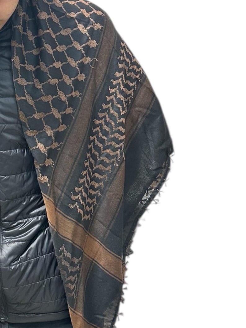 May include: A black and brown keffiyeh scarf with geometric and chevron patterns. The scarf has a fringed edge and is draped over a black jacket. The scarf's design features a combination of dark and light brown hues.