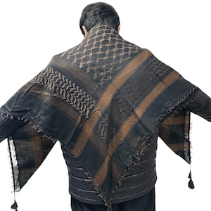 May include: Black and brown keffiyeh scarf with a geometric pattern and tassel trim. The scarf is draped over the shoulders. The scarf has brown stripes and a repeating pattern.
